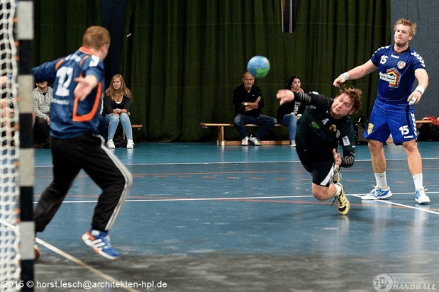 Zwei Handballspieler im Wettkampf, ein Spieler wirft den Ball auf das Tor, Zuschauer sitzen im Hintergrund.