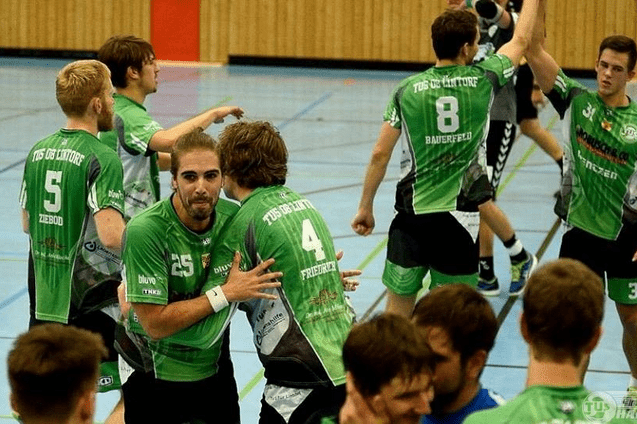 F&uuml;nf Handballspieler in gr&uuml;nen Trikots feiern auf einem Hallenboden mit Linien und schweren Ger&auml;ten im Hintergrund.