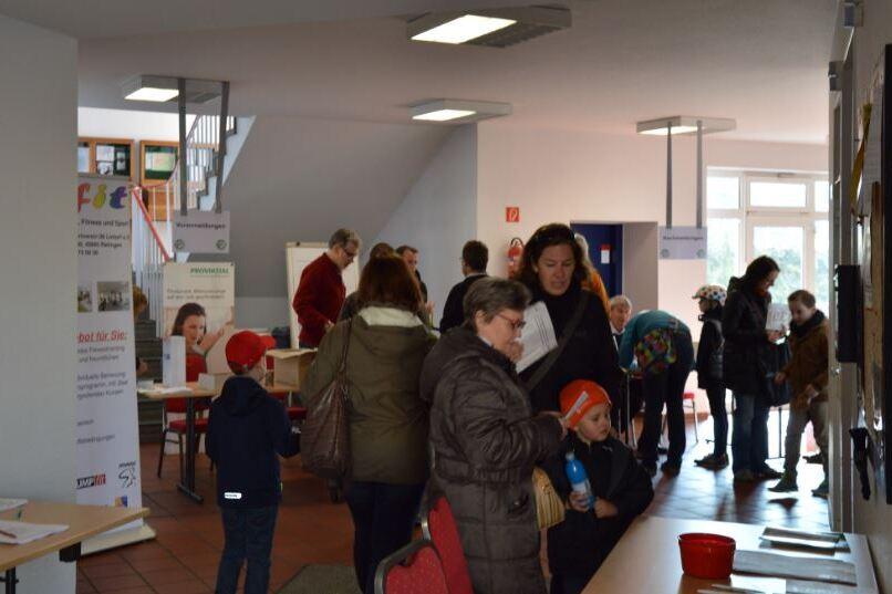 Menschen besprechen sich in einem &ouml;ffentlichen Raum mit Informationsst&auml;nden und Leseecke, einige halten Getr&auml;nke.
