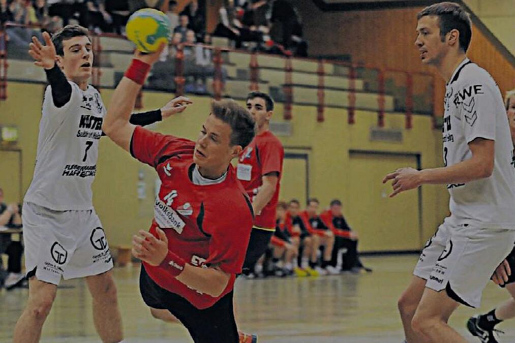 Spielszene beim Handball: Ein Spieler in Rot wirft den Ball, während zwei Verteidiger in Weiß ihn anfeuern.