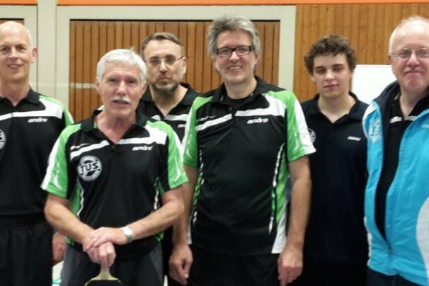 Sechs M&auml;nner in Tischtennis-Trainingsanz&uuml;gen posieren l&auml;chelnd in einer Sporthalle.