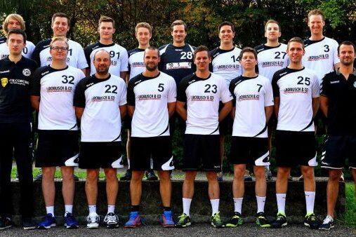 Mannschaftsbild von 15 Handballspielern in wei&szlig;en und schwarzen Trikots, auf einer Wiese vor B&auml;umen aufgestellt.