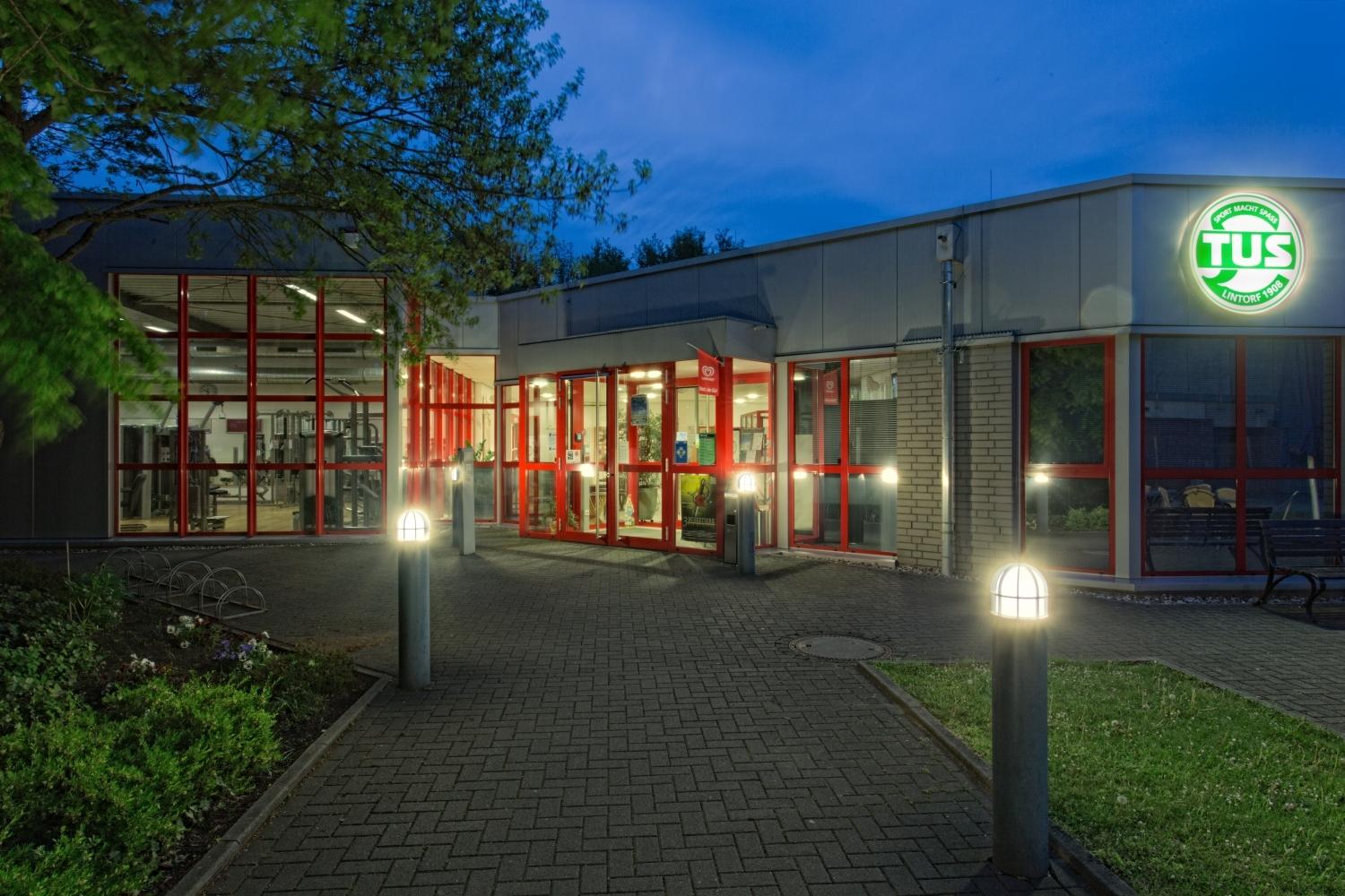 Eingang eines Sportzentrums mit roten Fenstern und beleuchteter TUS-Markierung, umgeben von bepflanzten Wegen bei Dämmerung.