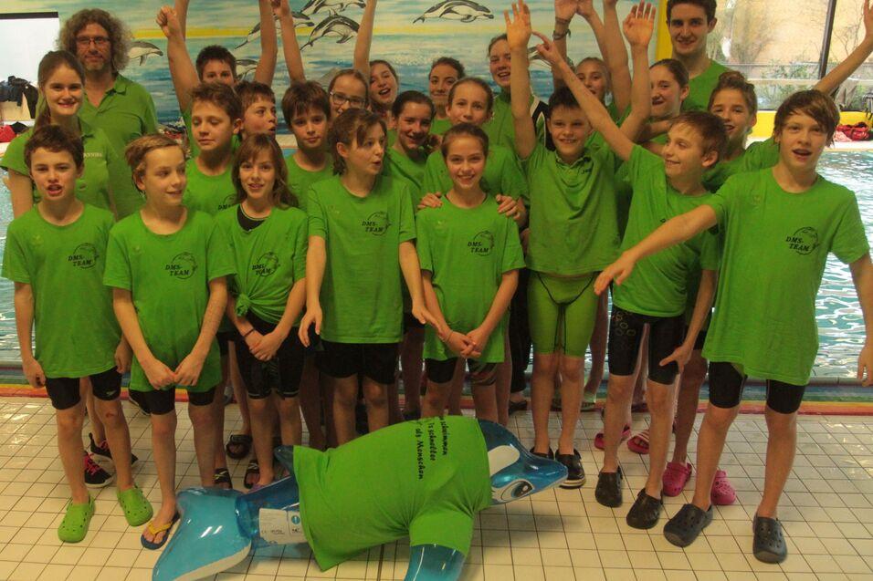 Gruppe von Kindern in gr&uuml;nen T-Shirts posiert l&auml;chelnd am Rand eines Schwimmbeckens mit einem aufblasbaren Delfin.
