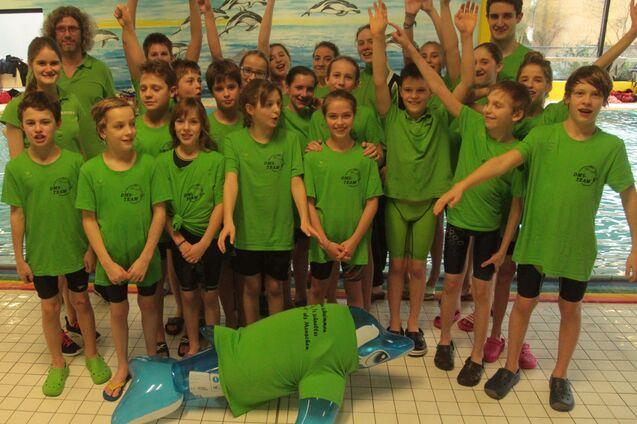 Gruppe von Kindern in gr&uuml;nen T-Shirts posiert l&auml;chelnd am Rand eines Schwimmbeckens mit einem aufblasbaren Delfin.