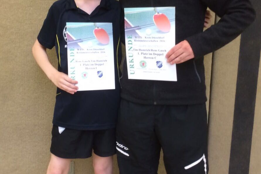 Zwei junge M&auml;nner stehen nebeneinander und halten jeweils eine Urkunde in der Hand, vor einer Wand in einer Sporthalle.