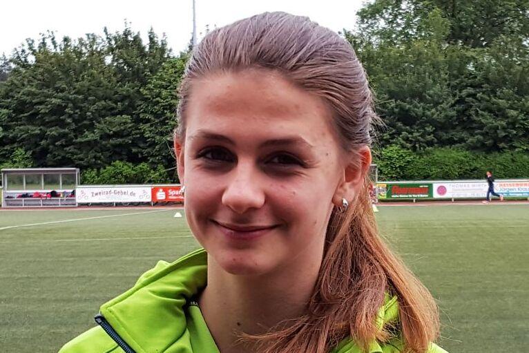 Portr&auml;t eines l&auml;chelnden M&auml;dchens in einer gr&uuml;nen Jacke vor einem Sportplatz mit Kunstrasen.