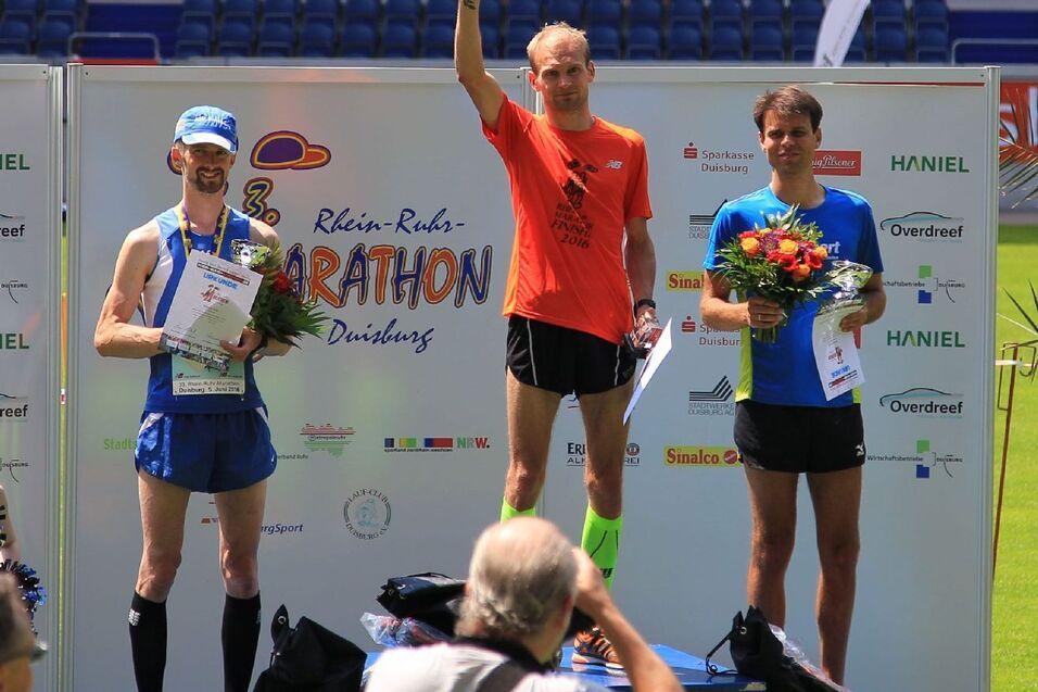 Drei Medaillengewinner stehen auf einem Podium bei einem Marathon, h&auml;lt Blumen und Urkunden in der Hand.