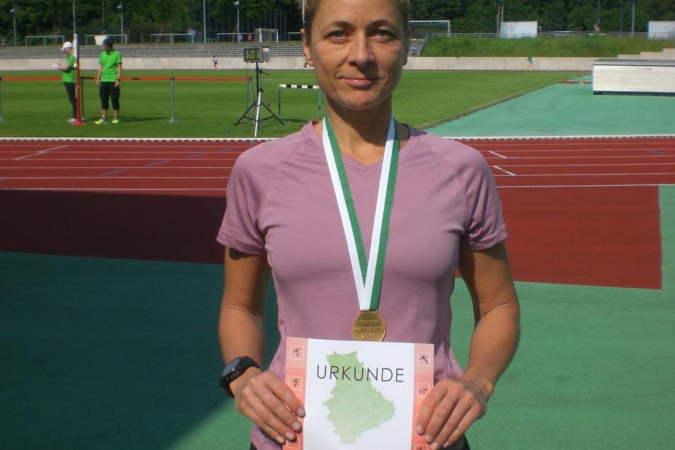 Athletin h&auml;lt stolz eine Urkunde und Medaille in der Hand, auf einem Sportplatz mit gr&uuml;ner Laufbahn im Hintergrund.