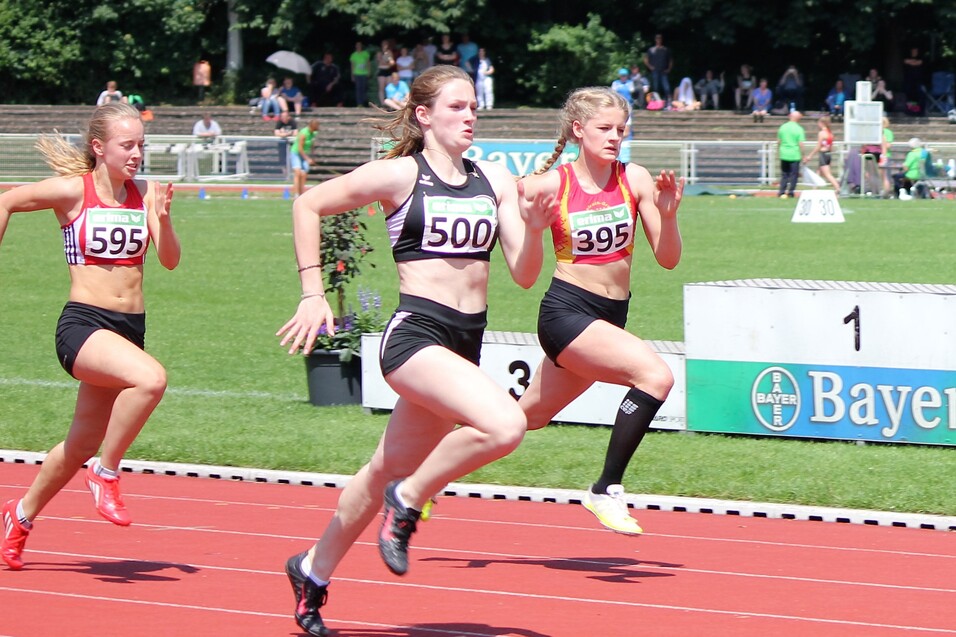 Drei athletische L&auml;uferinnen sprinten auf einer roten Laufbahn w&auml;hrend eines Wettkampfs im Freien.
