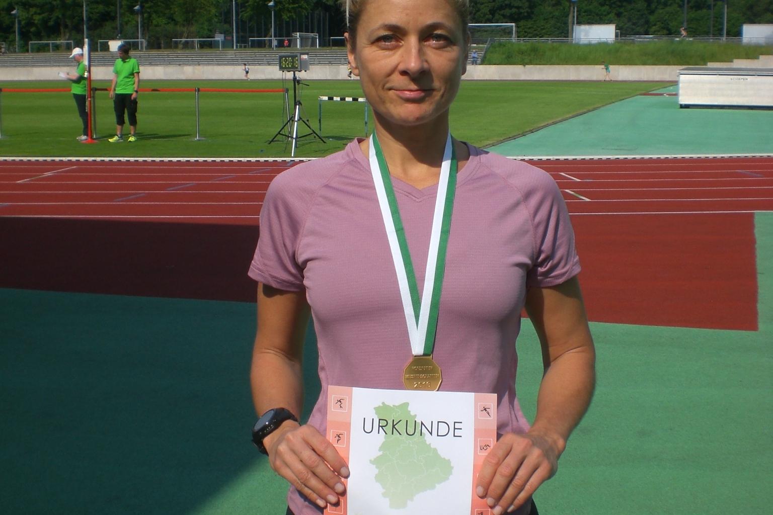 Athletin hält stolz eine Urkunde und Medaille in der Hand, auf einem Sportplatz mit grüner Laufbahn im Hintergrund.