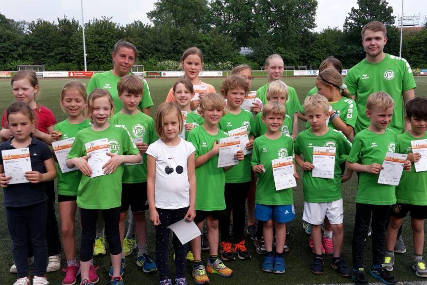 Gruppe von Kindern in gr&uuml;nen T-Shirts, l&auml;chelnd mit Urkunden, auf einem Fu&szlig;ballplatz hinter ihnen B&auml;ume und ein Sportfeld.