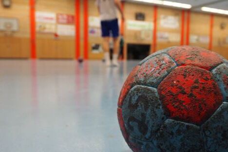 Nahaufnahme eines abgenutzten Handballs auf dem hallenblauen Boden, mit verschwommenem Spieler im Hintergrund.