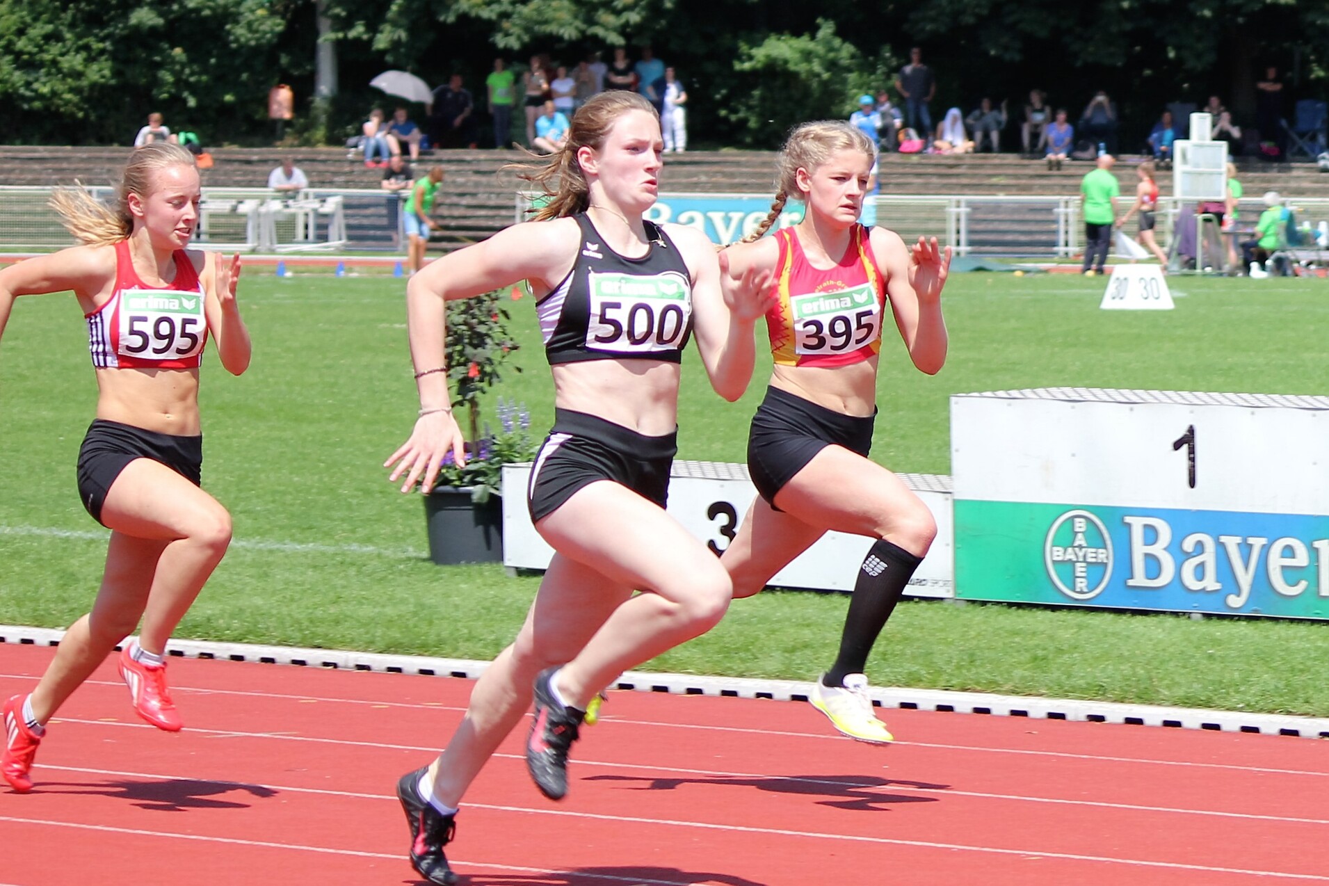 Drei athletische L&auml;uferinnen sprinten auf einer roten Laufbahn w&auml;hrend eines Wettkampfs im Freien.