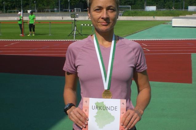 Athletin h&auml;lt stolz eine Urkunde und Medaille in der Hand, auf einem Sportplatz mit gr&uuml;ner Laufbahn im Hintergrund.