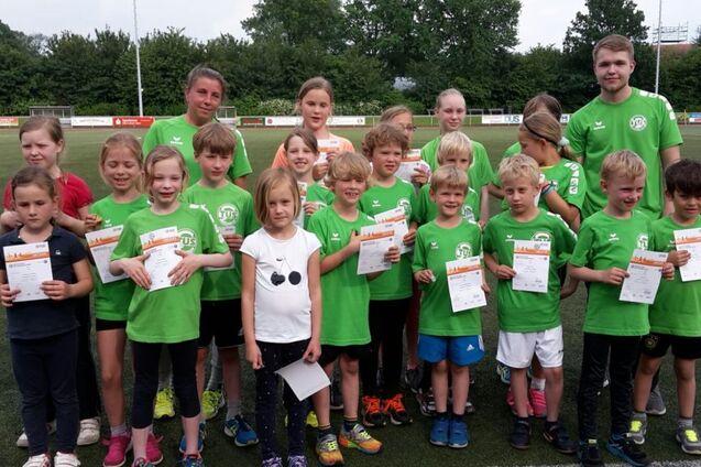 Gruppe von Kindern in gr&uuml;nen T-Shirts, l&auml;chelnd mit Urkunden, auf einem Fu&szlig;ballplatz hinter ihnen B&auml;ume und ein Sportfeld.