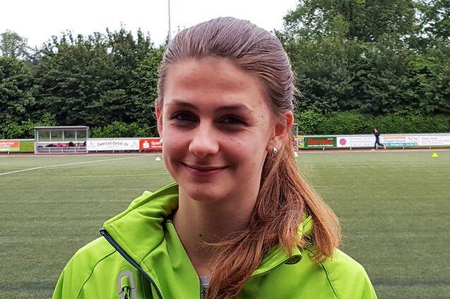 Portr&auml;t eines l&auml;chelnden M&auml;dchens in einer gr&uuml;nen Jacke vor einem Sportplatz mit Kunstrasen.