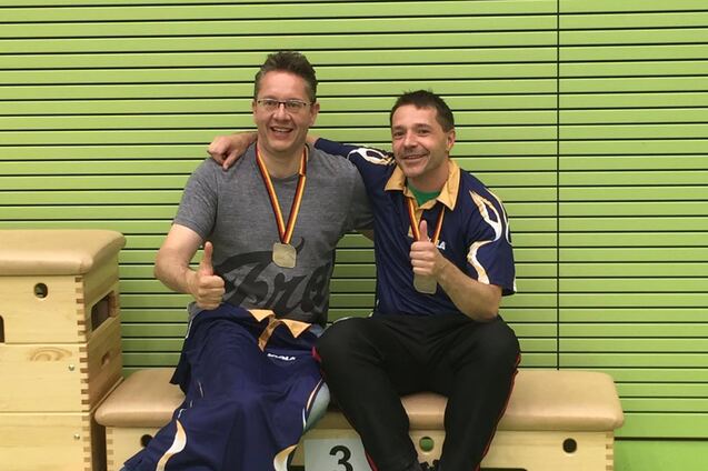 Zwei l&auml;chelnde M&auml;nner mit Medaillen sitzen auf einer Bank in einer Sporthalle und geben einen Daumen hoch.