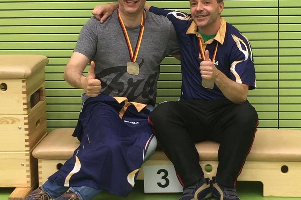 Zwei l&auml;chelnde M&auml;nner mit Medaillen sitzen auf einer Bank in einer Sporthalle und geben einen Daumen hoch.