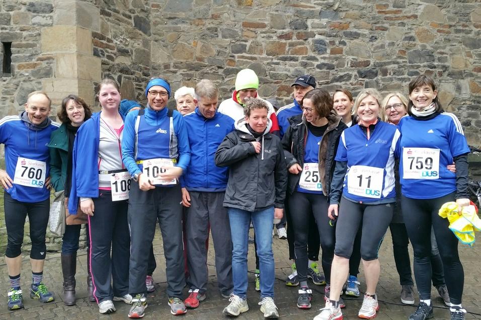 Gruppe von zw&ouml;lf L&auml;ufern in blauen Trikots mit Startnummern, posieren vor einem steinernen Hintergrund.
