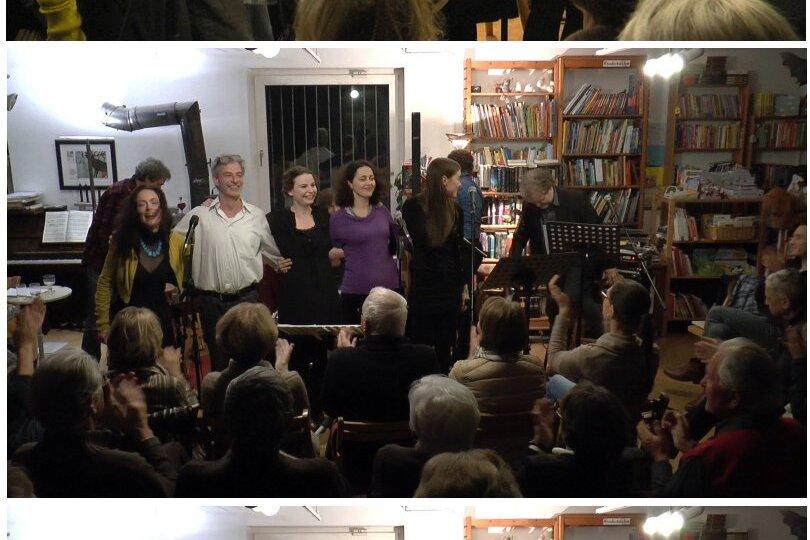 Fünf Sängerinnen und ein Musiker stehen auf einer kleinen Bühne, während das Publikum applaudiert. Bücherregale im Hintergrund.