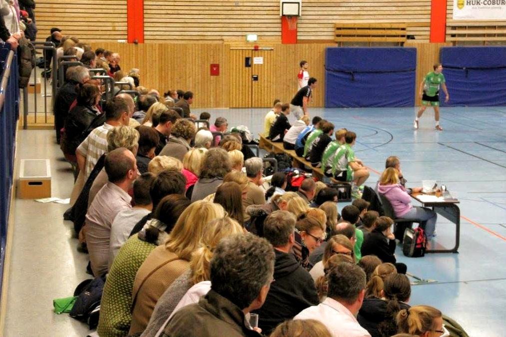 Zuschauer sitzen dicht gedrängt auf den Bänken in einer Sporthalle während eines Handballspiels.