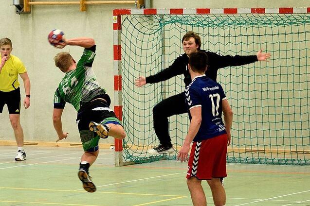 Handballspieler springt zum Wurf auf das Tor, w&auml;hrend ein Torwart sich zur Abwehr bereit macht und Schiedsrichter zuschaut.