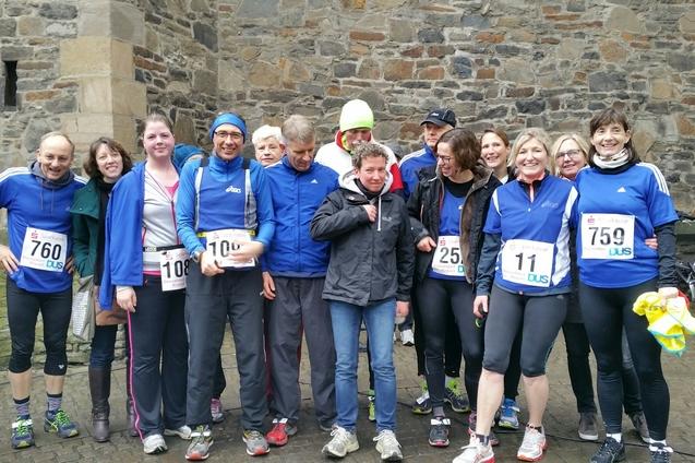 Gruppe von zw&ouml;lf L&auml;ufern in blauen Trikots mit Startnummern, posieren vor einem steinernen Hintergrund.