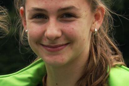 Nahaufnahme eines l&auml;chelnden M&auml;dchens mit langen, braunen Haaren in einer gr&uuml;nen Sportjacke, vor verschwommener Hintergrundlandschaft.