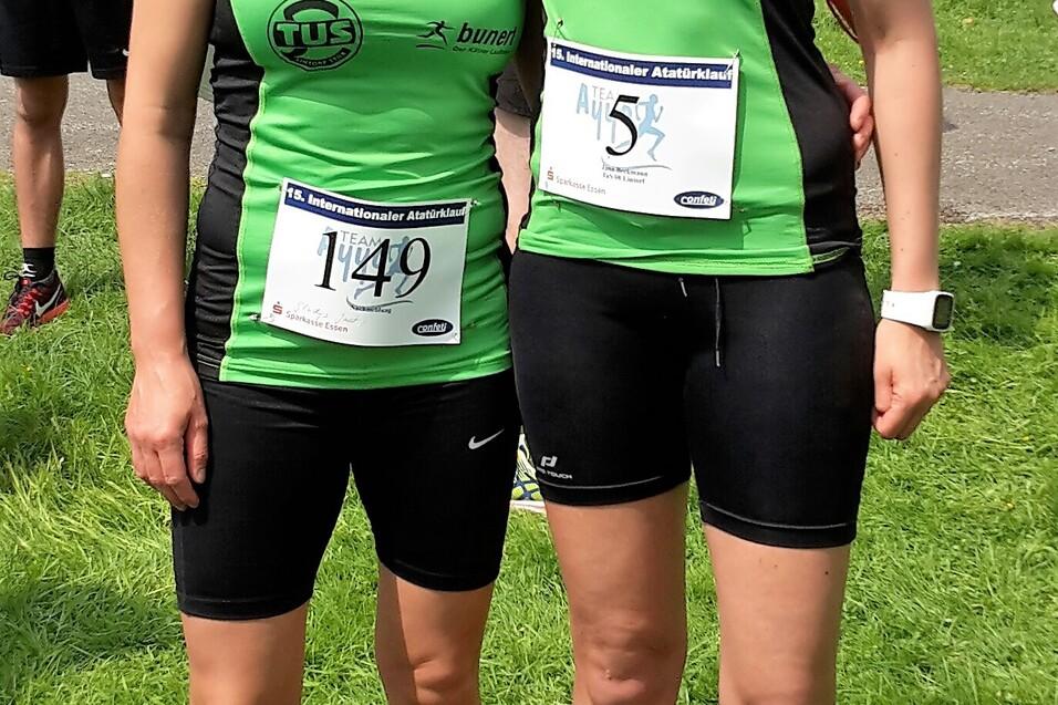 Zwei Athletinnen in gr&uuml;nen Laufshirts mit Startnummern stehen l&auml;chelnd auf einer Wiese vor einem Veranstaltungshintergrund.