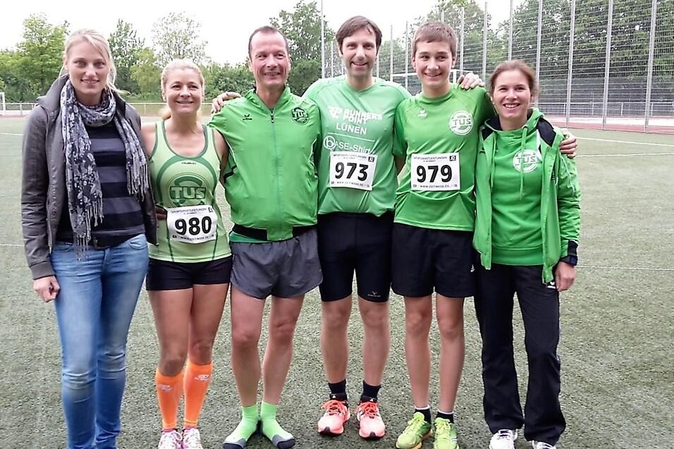 Sechs Personen in gr&uuml;ner Sportkleidung stehen auf einem Sportplatz, die meisten tragen Startnummern.