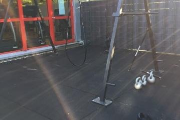 Fitnessbereich mit einem Trainingsgerät, einer Klimmzugstange und zwei Kettlebells auf rutschfester Fläche.