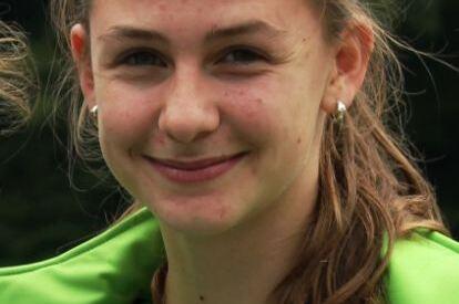 Nahaufnahme eines l&auml;chelnden M&auml;dchens mit langen, braunen Haaren in einer gr&uuml;nen Sportjacke, vor verschwommener Hintergrundlandschaft.