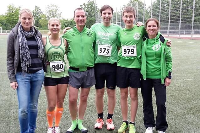 Sechs Personen in gr&uuml;ner Sportkleidung stehen auf einem Sportplatz, die meisten tragen Startnummern.