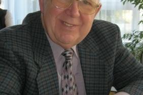 &Auml;lterer Mann mit Brille in kariertem Anzug, l&auml;chelt am Tisch, umgeben von Pflanzen und Tageslicht.