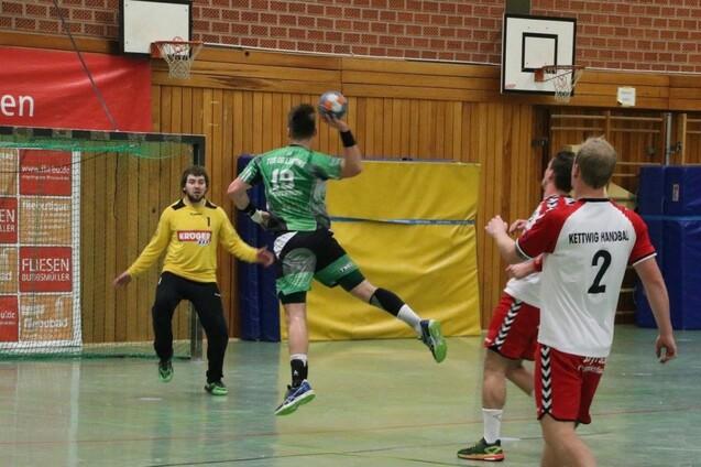 Handballspieler in gr&uuml;ner Uniform springt zum Tor und wirft den Ball, w&auml;hrend der Torwart in gelber Ausr&uuml;stung reagiert.