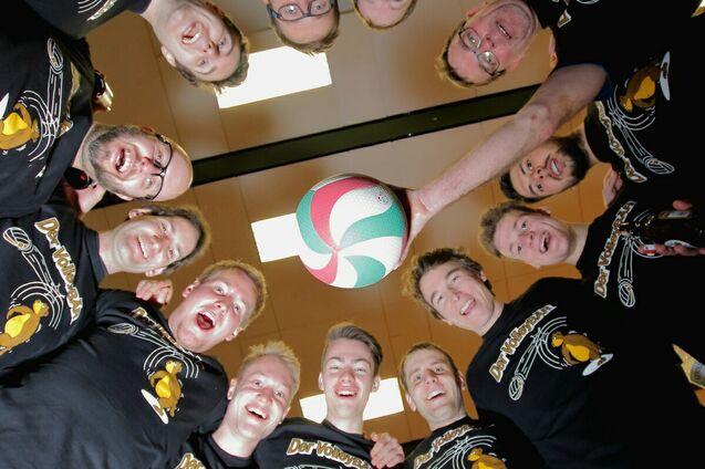 Gruppenselfie von zw&ouml;lf M&auml;nnern in schwarzen T-Shirts, die gemeinsam einen bunten Volleyball von oben halten.
