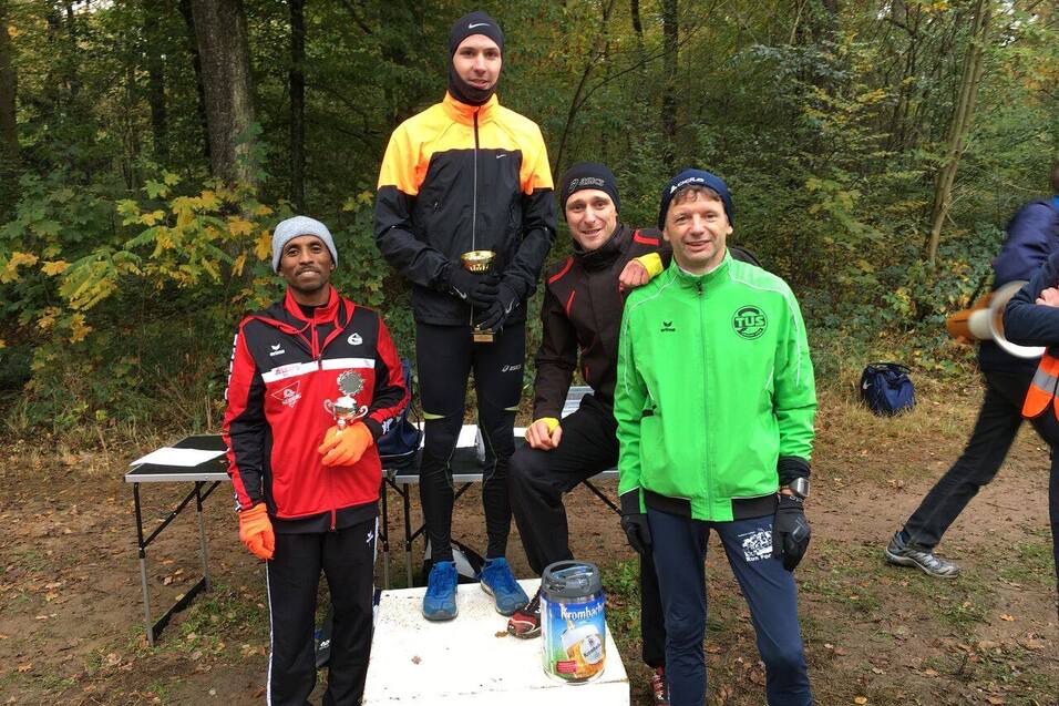 Vier L&auml;ufer stehen auf einem Podest im Wald, einer h&auml;lt einen Pokal, w&auml;hrend die anderen Medaillen zeigen.