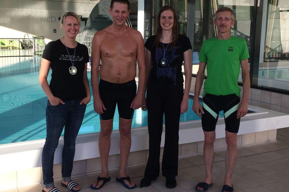 Vier Erwachsene stehen vor einem Schwimmbecken, einige tragen Schwimmbekleidung und Medaillen.