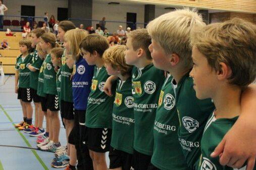 Gruppe von Kindern in Sporttrikots stehen in Reihe, einige halten sich an den Schultern, in einer Sporthalle.