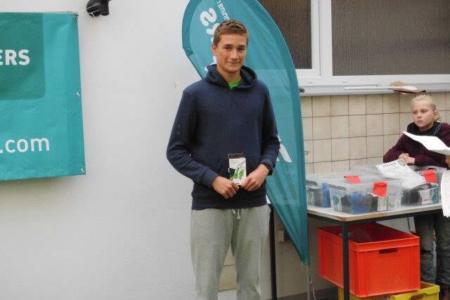 Junger Mann mit Hoodie hält eine Auszeichnung, steht vor einem Banner und einem Tisch mit Boxen in einem Innenraum.