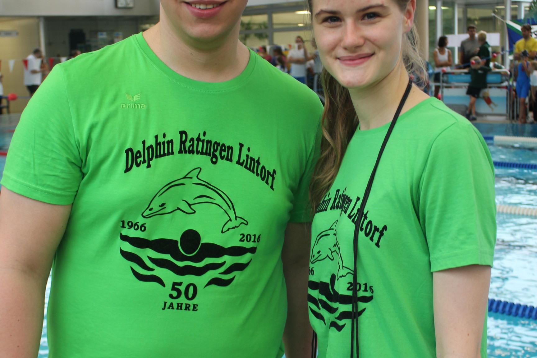Zwei Personen in grünen T-Shirts mit Delphin-Motiv stehen am Beckenrand eines Schwimmbads.