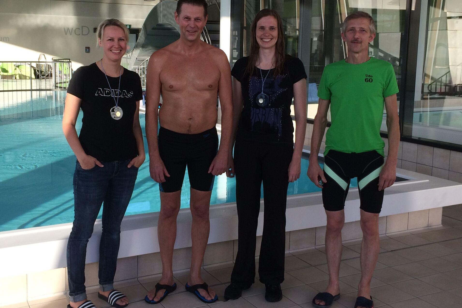 Vier Erwachsene stehen vor einem Schwimmbecken, einige tragen Schwimmbekleidung und Medaillen.