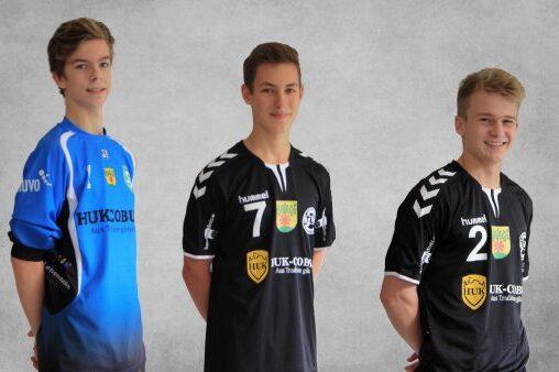 Drei junge Handballspieler posieren in unterschiedlichen Trikots, zwei in schwarz, einer in blau, vor einem grauen Hintergrund.
