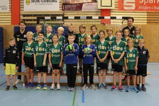 Gruppensicht von Kindern in gr&uuml;nen Handballtrikots, vor einer Sporthalle, einige halten Handb&auml;lle, Trainer im Hintergrund.