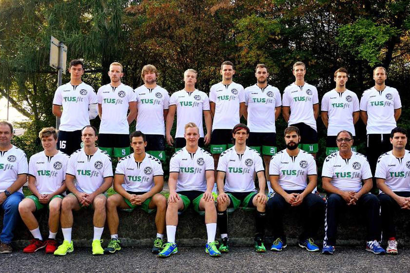 Gruppenfoto einer M&auml;nnermannschaft mit 15 Spielern in wei&szlig;en T-Shirts und gr&uuml;nen Hosen, auf einer Bank im Freien.
