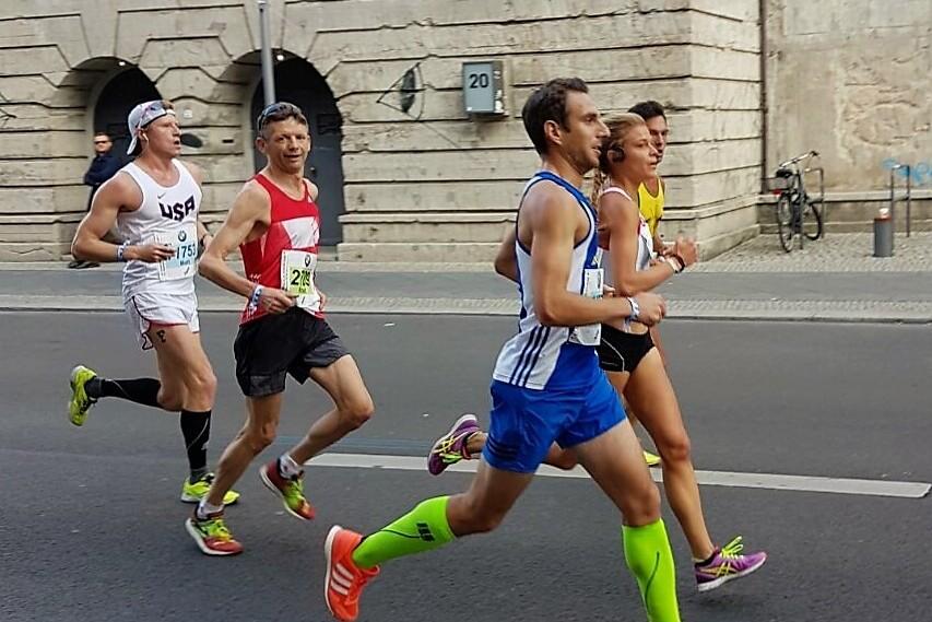 Vier L&auml;ufer in Wettkampfbekleidung joggen auf einer Stra&szlig;e mit historischen Geb&auml;uden im Hintergrund.