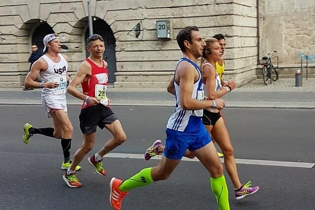 Vier L&auml;ufer in Wettkampfbekleidung joggen auf einer Stra&szlig;e mit historischen Geb&auml;uden im Hintergrund.