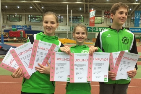 Drei Kinder in gr&uuml;nen Sportoutfits halten Urkunden in einer Sporthalle mit Laufbahn im Hintergrund.
