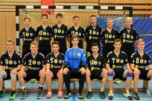 Mannschaftsfoto einer Handballmannschaft mit 14 Spielern in schwarzen Trikots und einem Torwart in Blau, vor einem Handballtor.